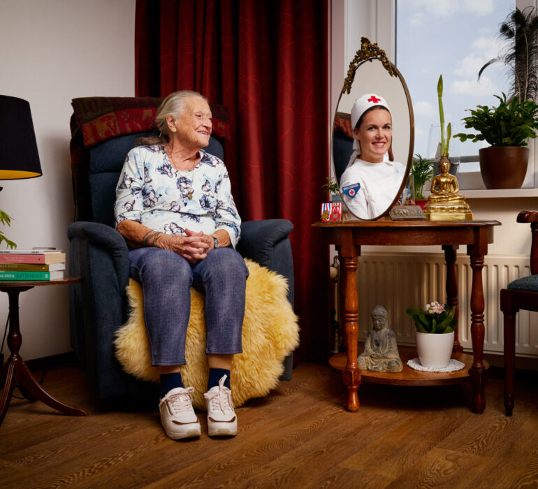 Mevrouw kijkt in de spiegel naar een jongere versie van haarzelf in verpleegkleding.