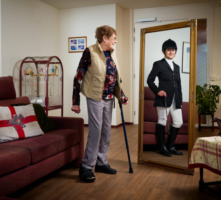 Mevrouw kijkt in de spiegel naar een jongere versie van haarzelf in paardrij kleding.